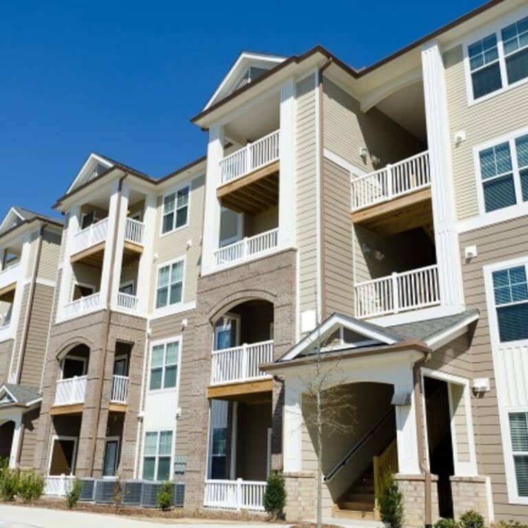 A three-story apartment complex with clean siding, white railings, and brick accents under a clear blue sky, showcasing the results of professional pressure washing for multi-family properties.