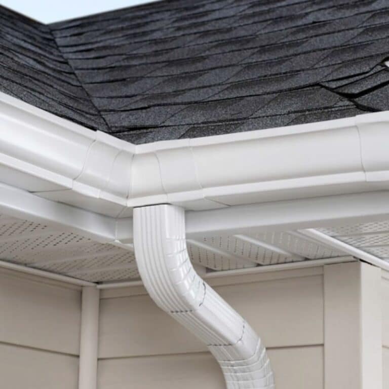 A close-up of a clean, white gutter system with downspout and leaf guard installed under a shingle roof, showcasing the results of professional gutter cleaning and protection services.
