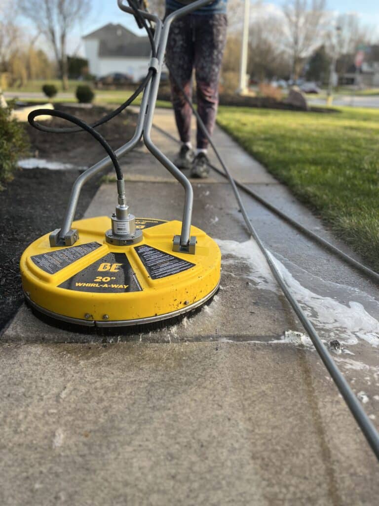 A technician uses a yellow surface cleaner to wash a concrete sidewalk, creating visible suds and a clean path, demonstrating effective pressure washing services in Perrysburg, OH.